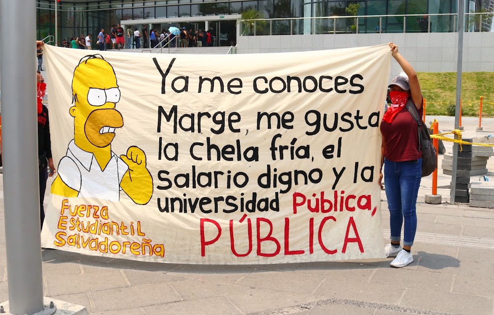 Person holding a FUerza Estudiantil Salvadorena banner that says "Ya me conoces Marge, me gusta la chela fria, el salario digno, y la universidad publica, PUBLICA"