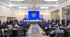 A big room with people sitting at desks and a state with the seal of El Salvador behind it.