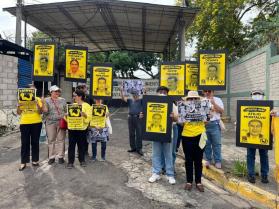 A dozen people stand in front of a gray gate  with signs that have names of political prisoners