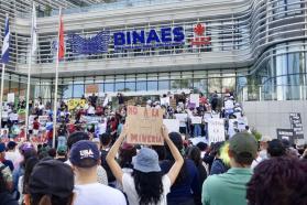 A person with long hair holds a sign saying "No a la minería" in front of a crowd gathered on the steps of a building. The building has a sign that says BINAES.