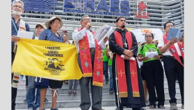 A line of peope holding a yellow flag the says "Liibertad para los lideres comunitarios" speaks to a crowd with the National Library in the background