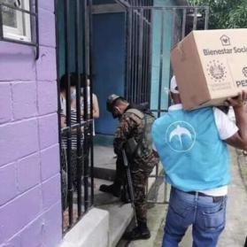 Two people stand in a doorway as one person wearing a military uniform approaches followed by another person with a teal vest with GANA (previously Nuevas Ideas logo) carrying a box marked with Salvadoran government logo.