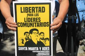 A person holding a yellow and black sign with the faces of five people that says "Libertad para los líderes comunitarios"