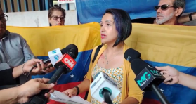 Marisela Ramirez stands in front of people holding Venezuelan flags, speaking into microphones held by journalists