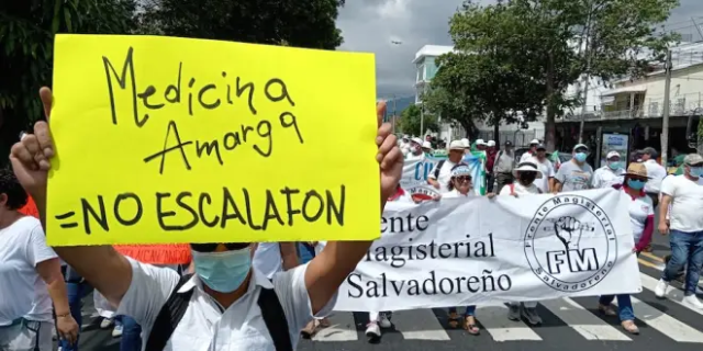 Man holds sign at march that reads, in Spanish, "Bitter medicine equals no pay scale" in front of a teachers union banner