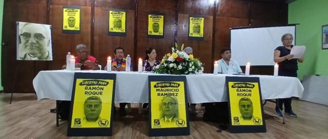 A panel of speakers sits at a table surrounded by poster of Mauricio Landaverde and Ramon Roque