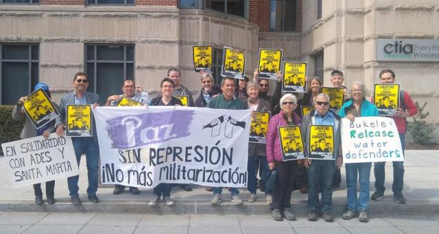 A group of 17 people with signs and a banner that says "Paz sin represion, No mas militarizacion" in front of a gray building