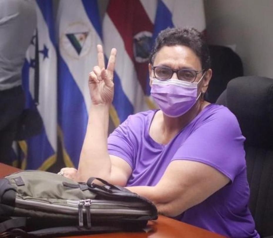 Photo of Lorena Pena wearing purple, sitting at table holding up a peace sign with two fingers